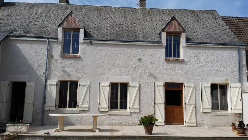 Façade en pierre d'une maison avec volets blancs, porte en bois ouverte et plantes en pot devant.