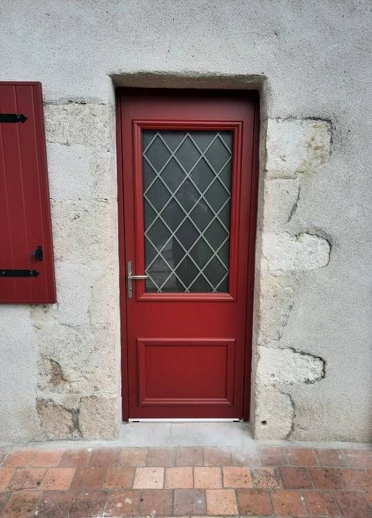 Porte rouge à vitrage en losanges encastrée dans un mur de pierre au-dessus d'un dallage.