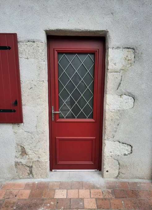 Porte rouge à vitraux en losanges dans un mur de pierre, à côté d'un volet rouge sur une allée carrelée.