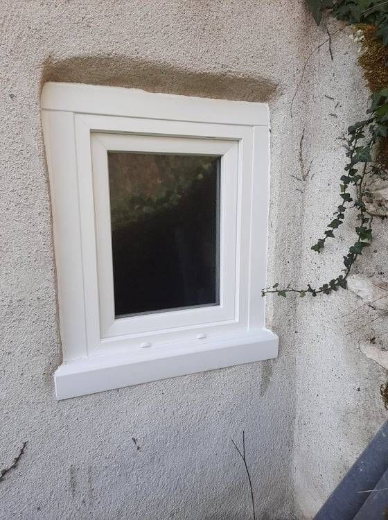 Petite fenêtre blanche encastrée dans un mur en stuc texturé, avec du lierre grimpant le long du côté droit.