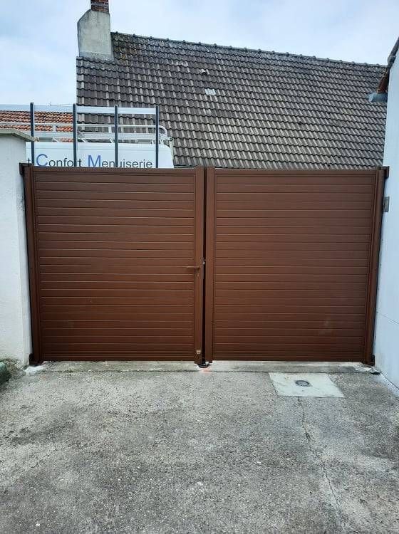 Un portail double marron bloque l'accès à une allée devant un bâtiment au toit de tuiles.