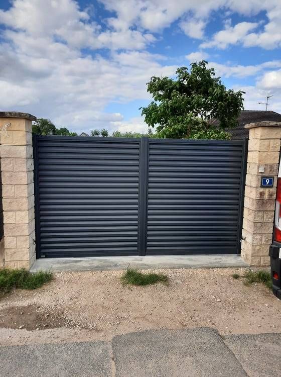 Portail en métal gris foncé entre des piliers de pierre sous un ciel partiellement nuageux