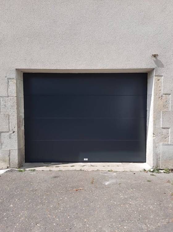 Porte de garage bleu foncé encastrée dans un mur de pierre claire, à côté d'une allée pavée.
