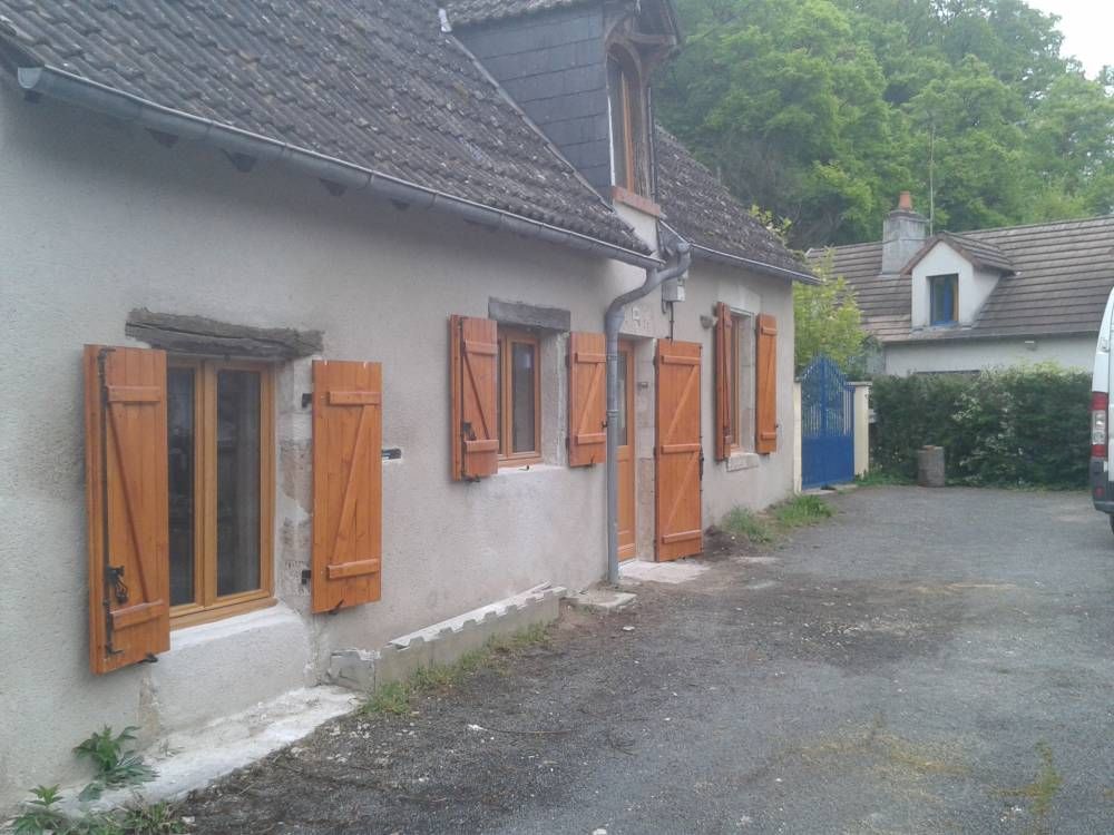 Extérieur d'une maison de campagne aux murs gris, volets en bois et allée de gravier sous un ciel nuageux.
