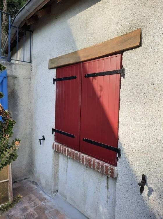 Volet en bois rouge sur un mur en stuc, avec des charnières noires.