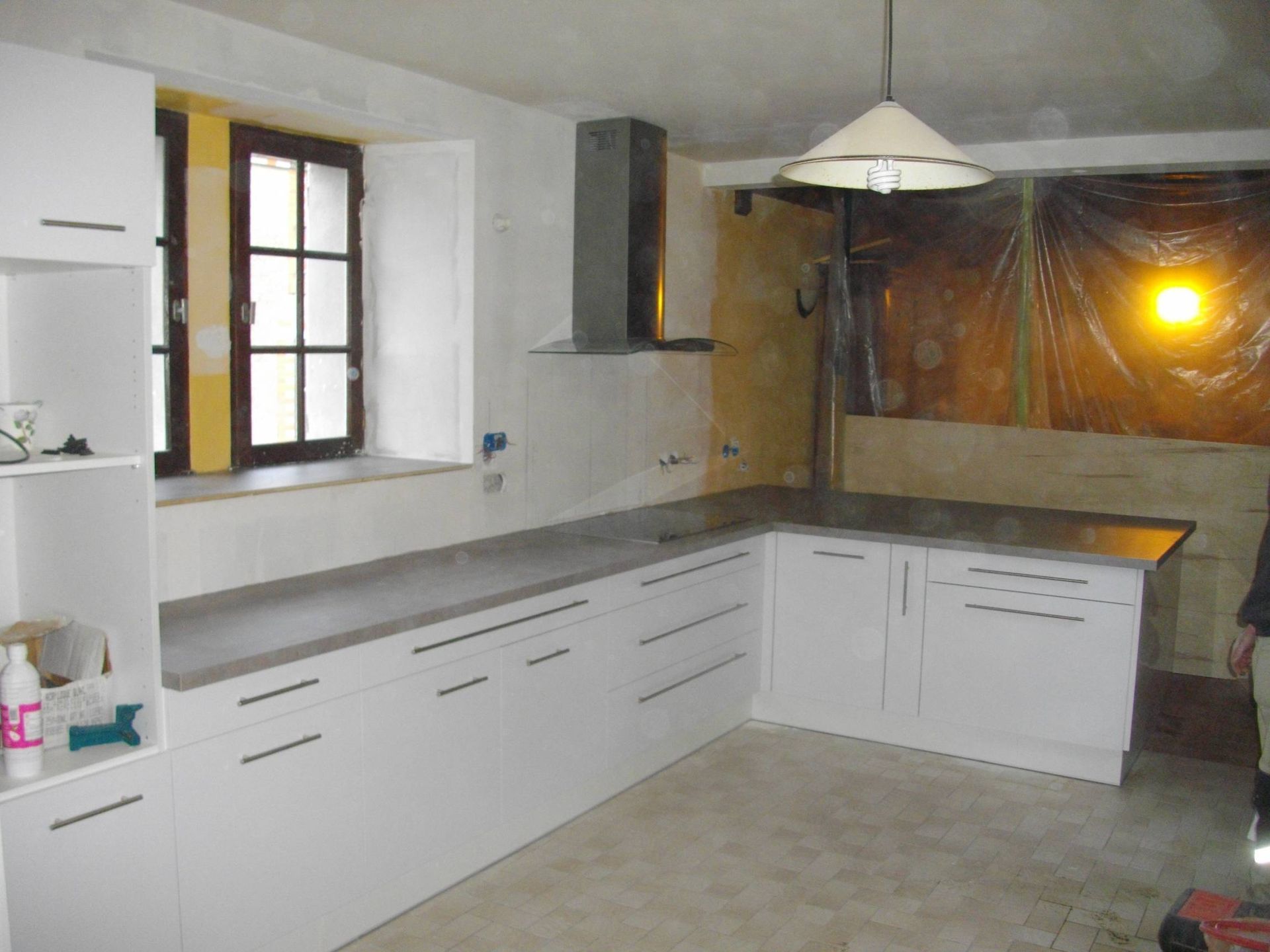 Cuisine blanche en forme de L avec placards, évier avec fenêtre, suspension lumineuse et crédence non finie.