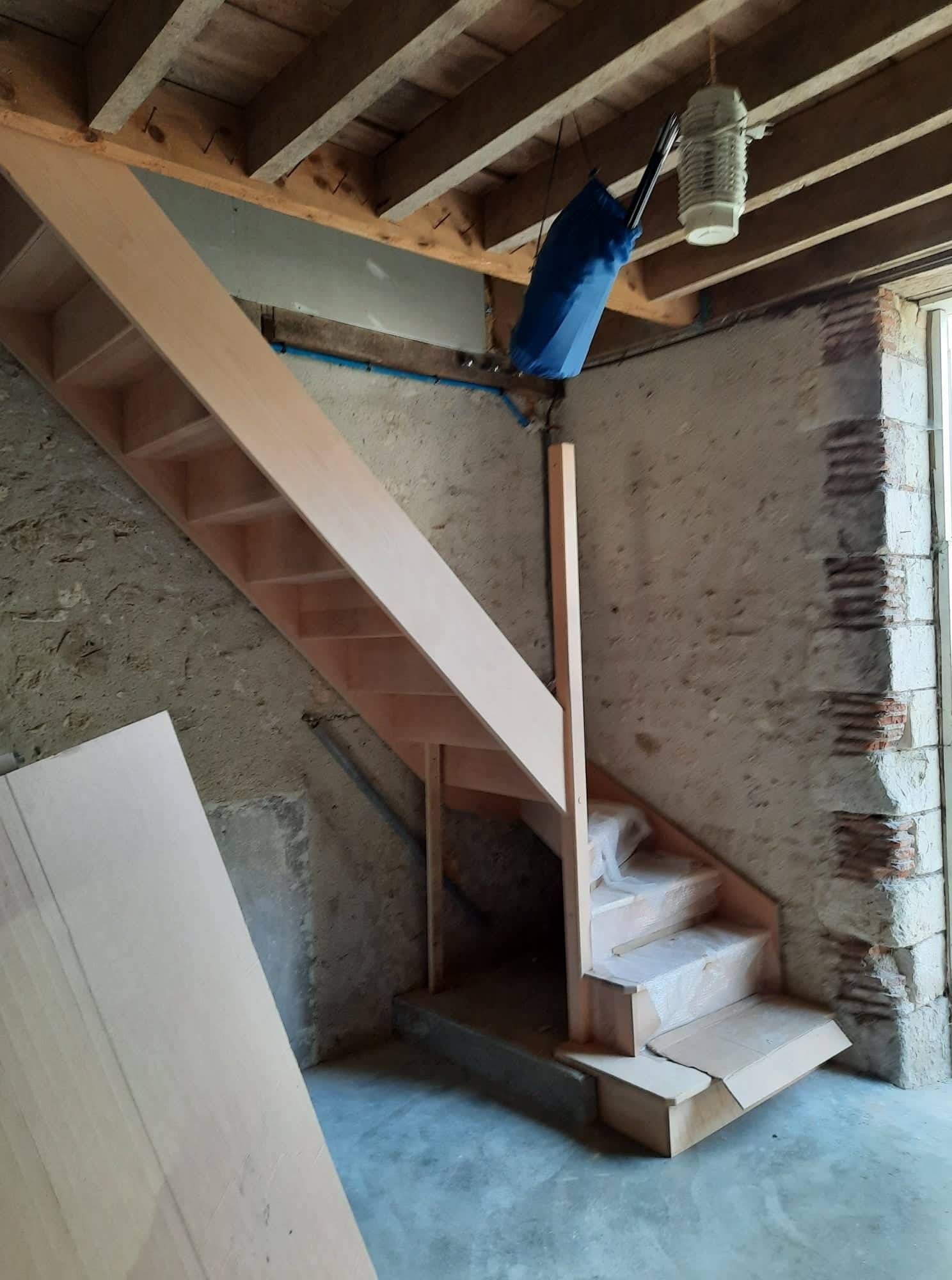 Escalier intérieur inachevé avec poutres en bois apparentes, mur en béton et luminaire suspendu