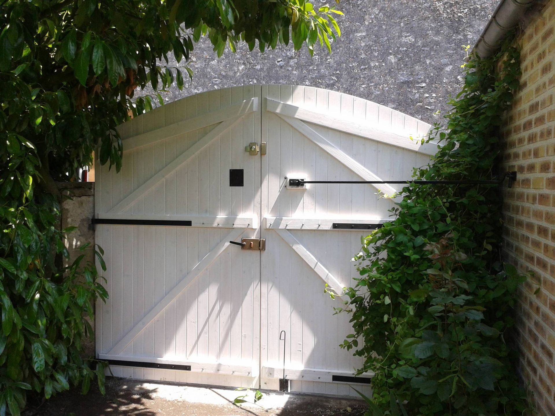 Portail de jardin en bois blanc encadré de murs de briques et d'une végétation dense baignée de soleil