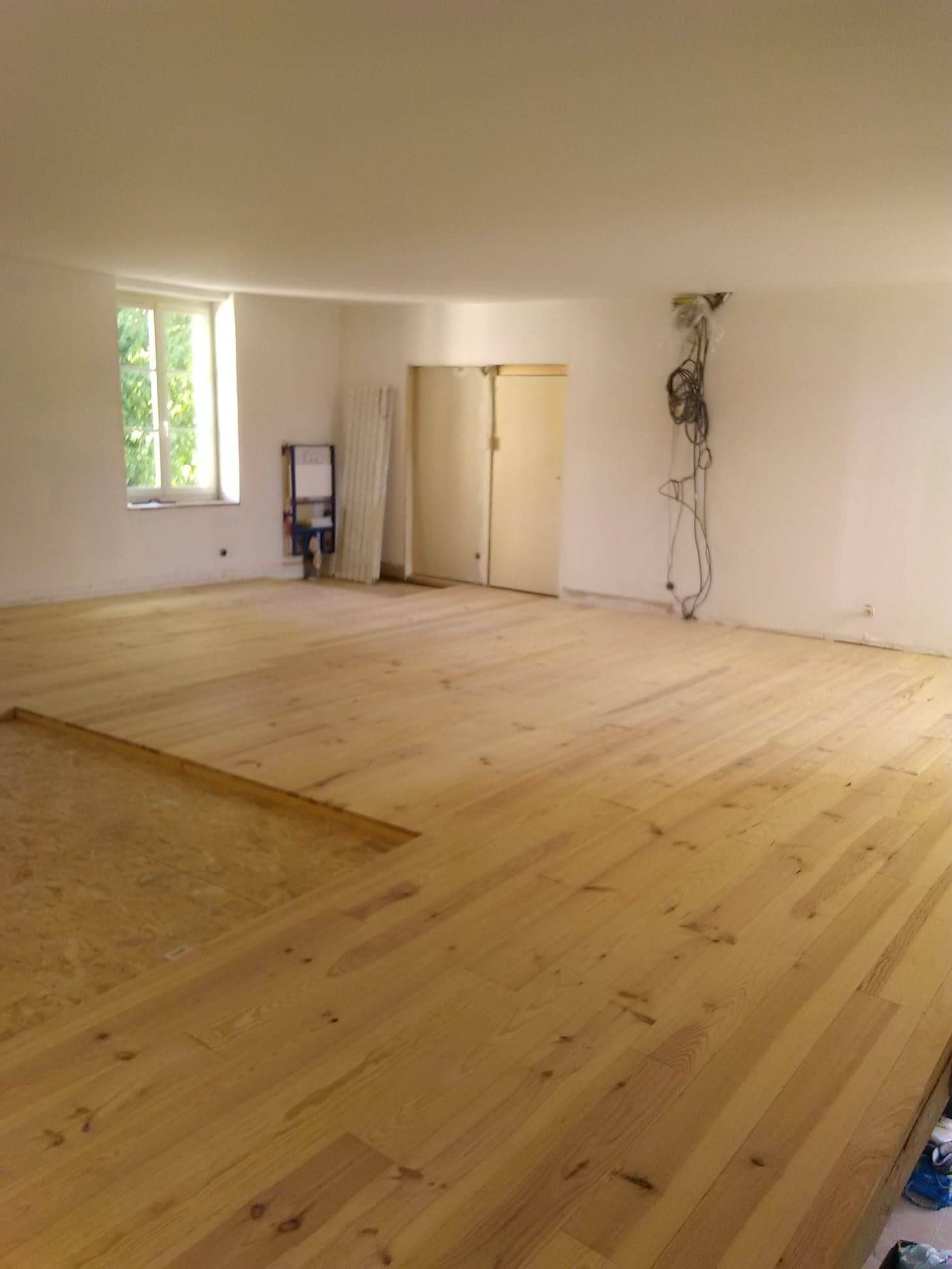 Pièce vide avec parquet clair, murs blancs, fenêtre et portes doubles ; câbles électriques apparents à droite.