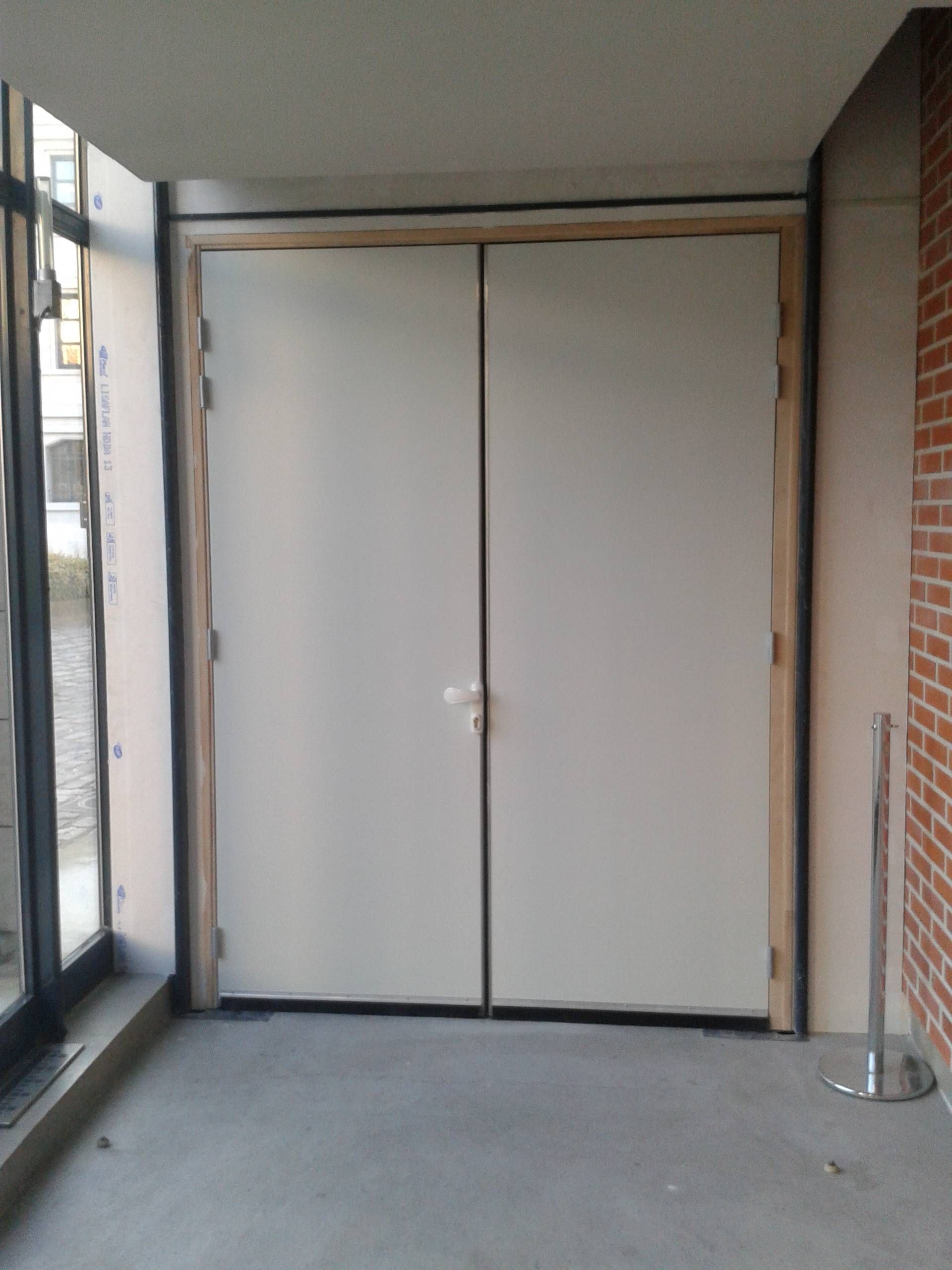 Double porte blanche fermée dans un couloir étroit avec de grandes fenêtres et des murs en briques.