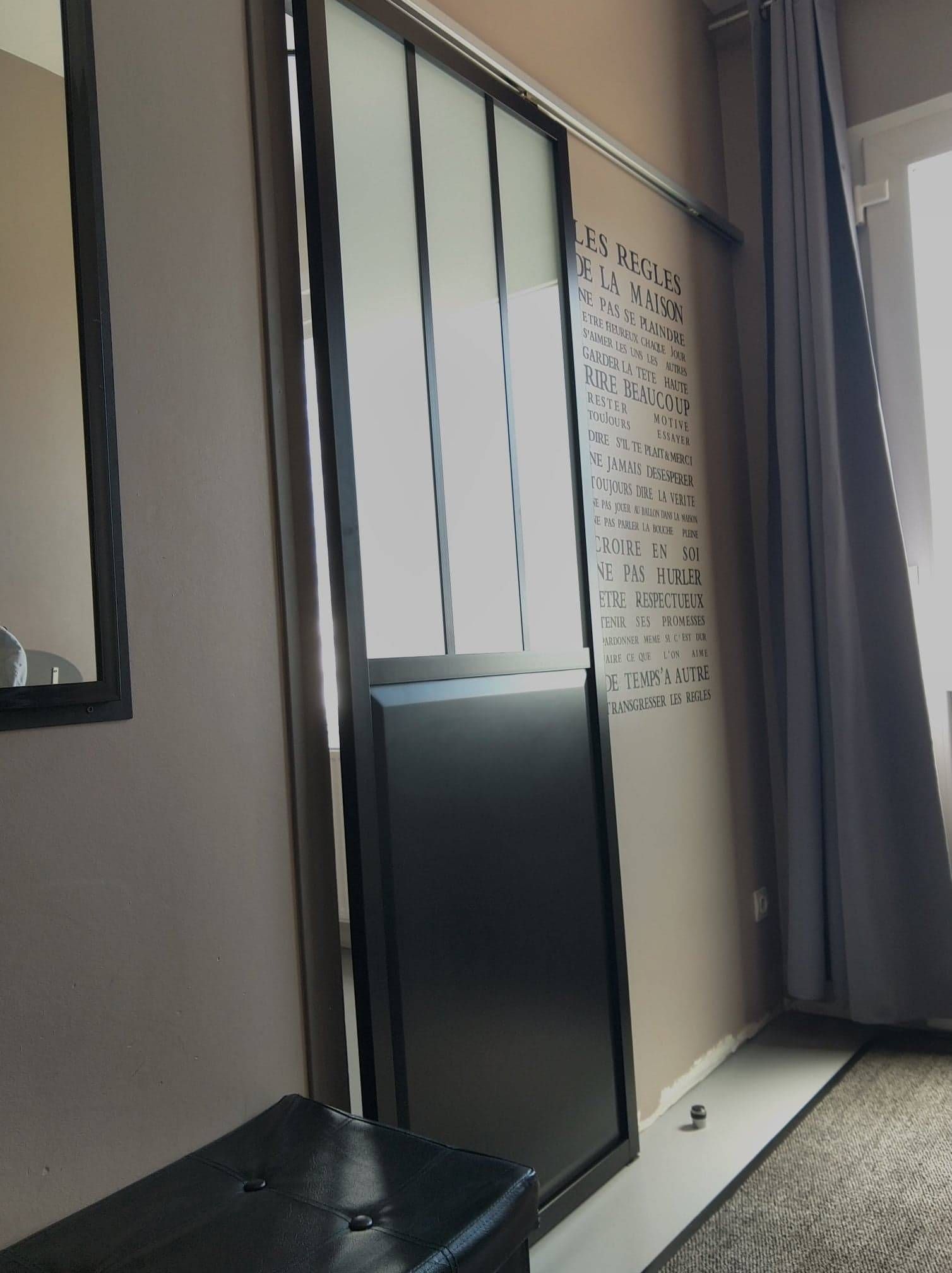 Pièce moderne avec de hautes fenêtres, des rideaux gris, un miroir et une armoire sombre près d'un panneau mural à motifs.