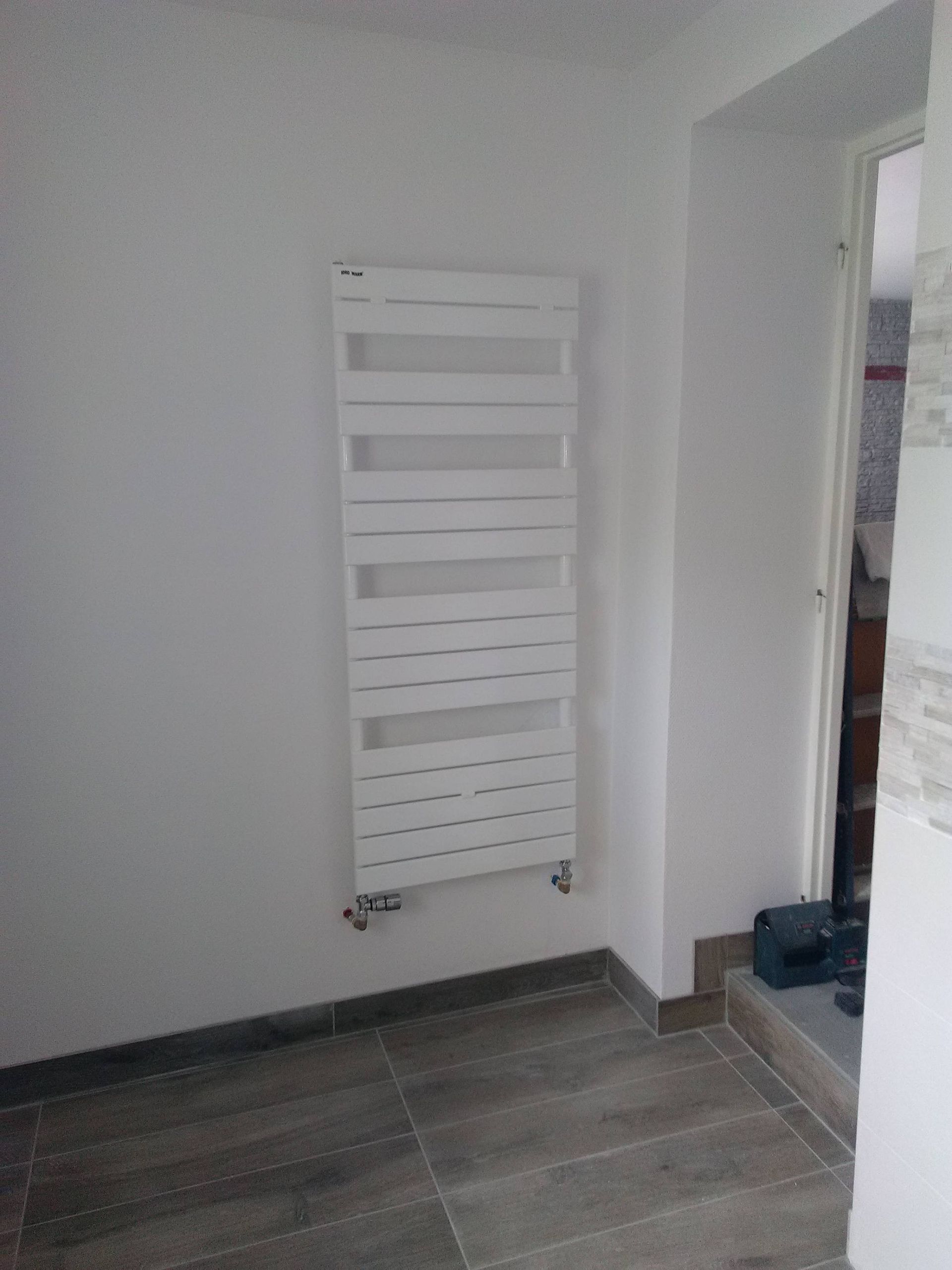 Radiateur mural blanc dans un couloir carrelé avec une porte ouverte sur la droite.