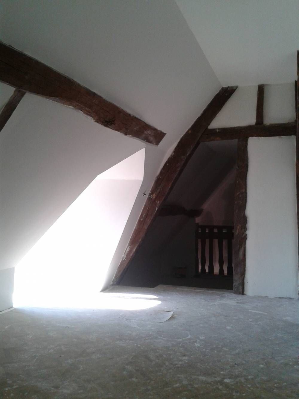 Grenier vide avec poutres en bois inclinées, murs blancs et lumière vive provenant d'une fenêtre basse.