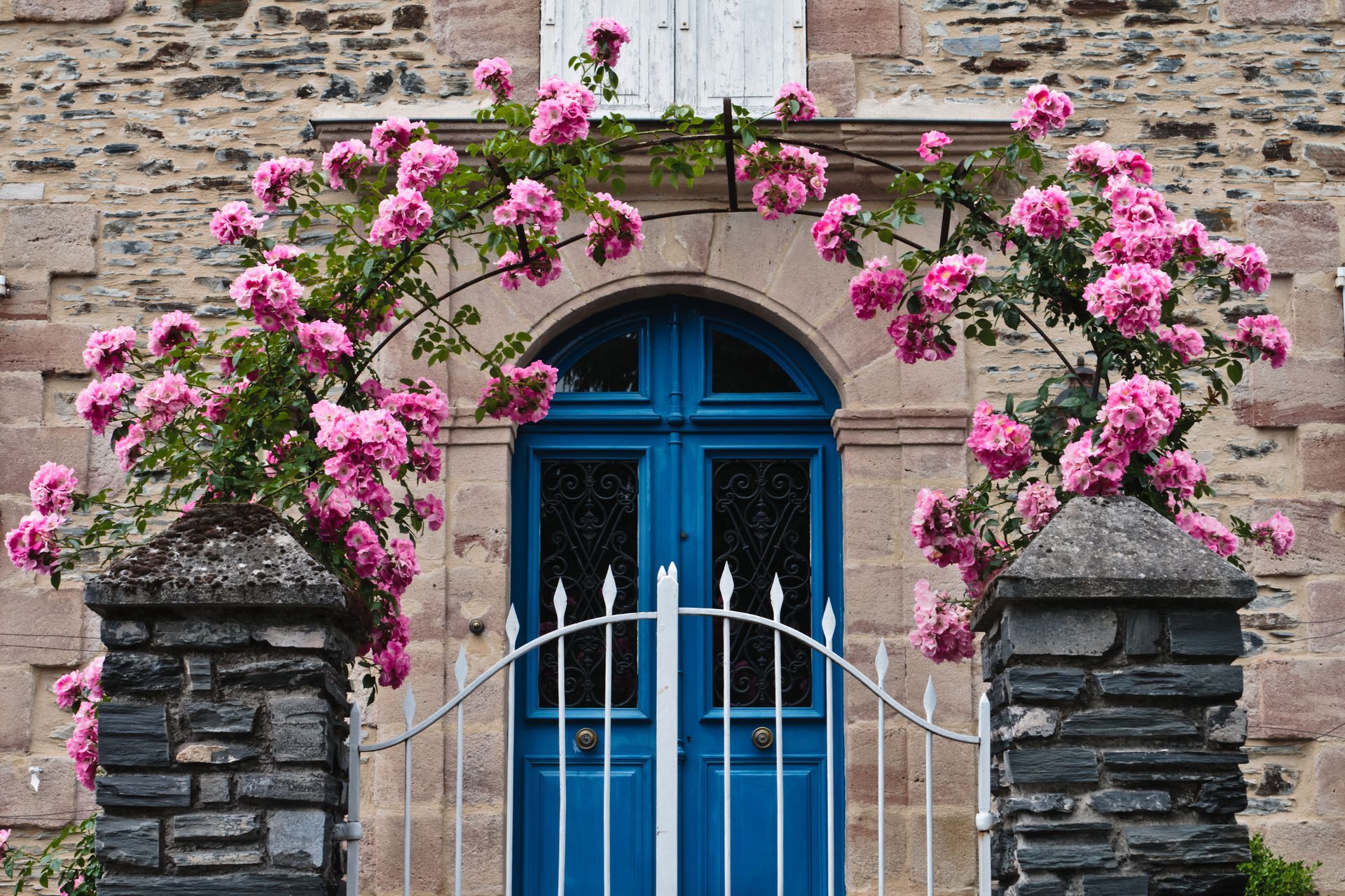 Porte cintrée bleue avec portail blanc, encadrée de fleurs roses sur une façade de maison en pierre
