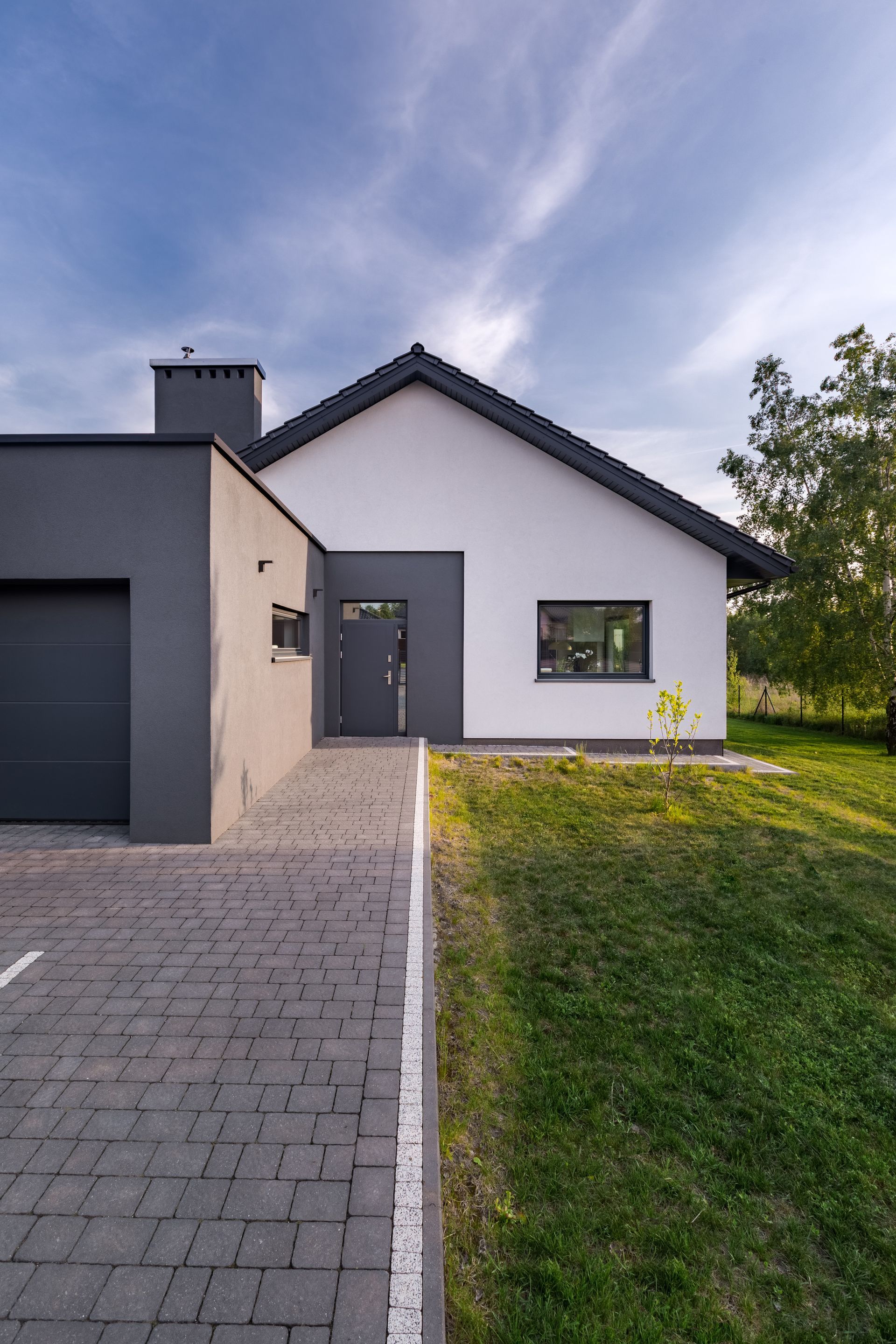 Maison blanche moderne avec des finitions grises, une allée, un garage et une pelouse à l'avant sous un ciel nuageux
