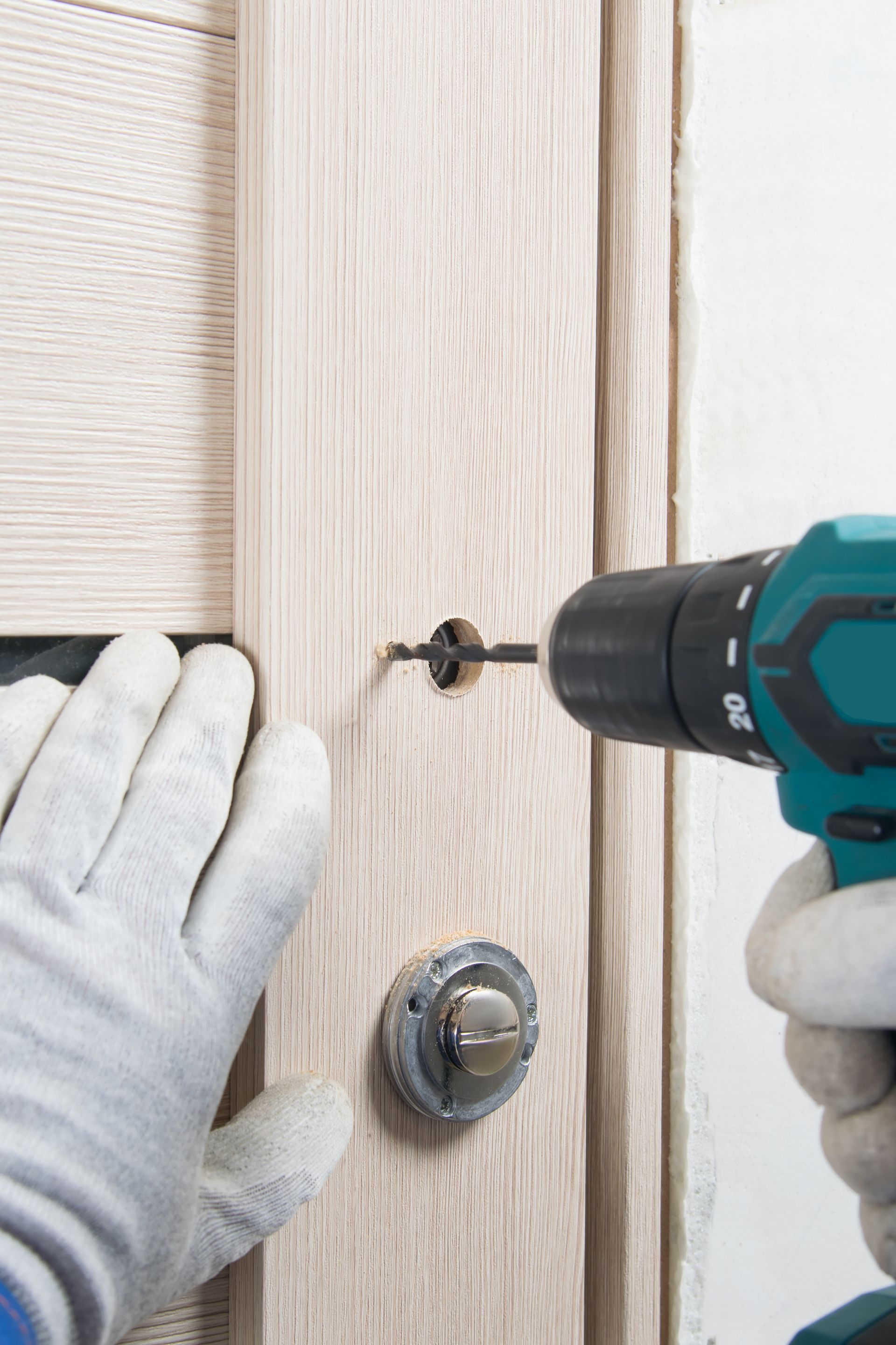 Des mains gantées utilisent une perceuse pour installer une serrure sur une porte en bois clair