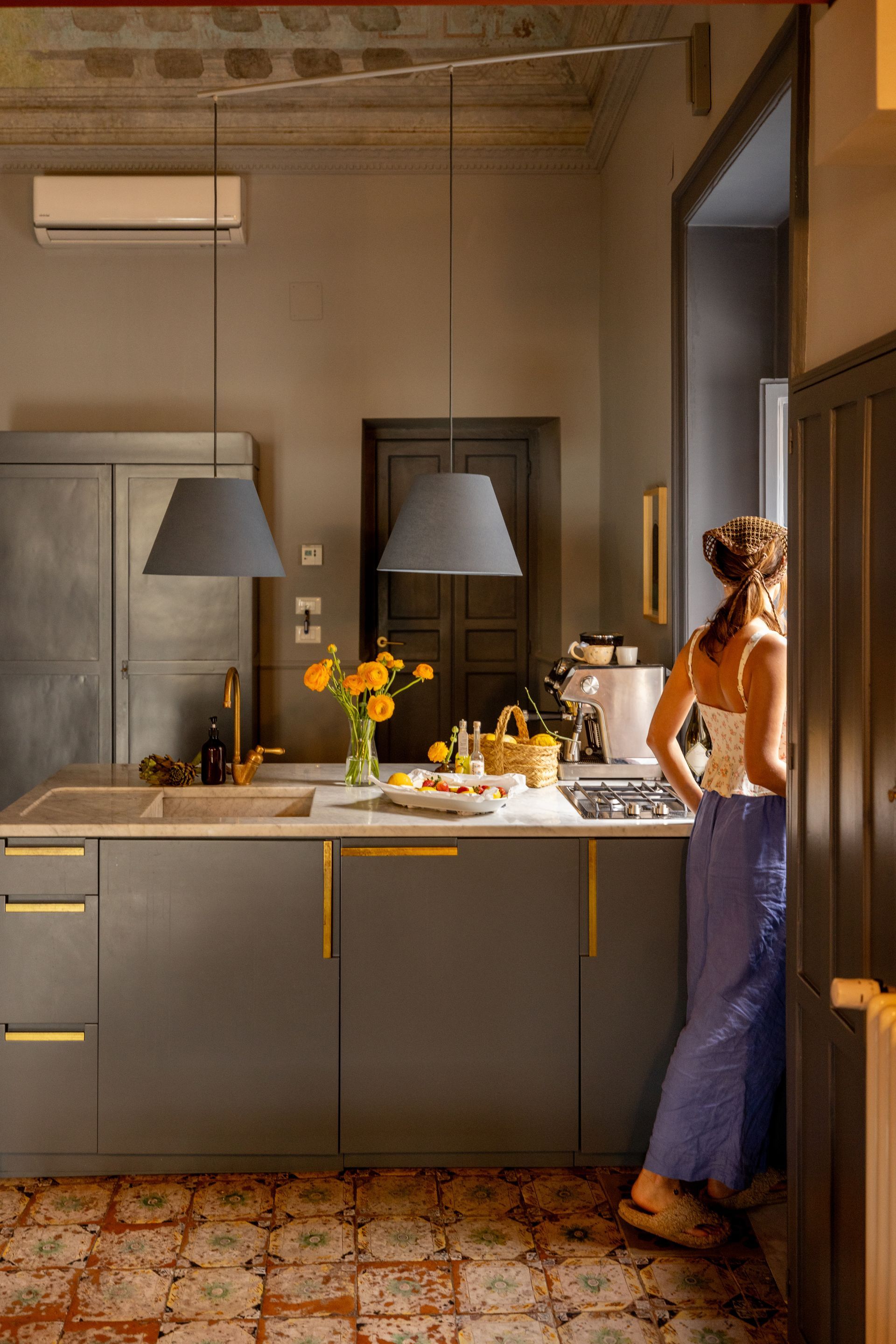 Cuisine moderne avec placards gris, suspensions lumineuses, vase de fleurs et une personne debout près du comptoir.