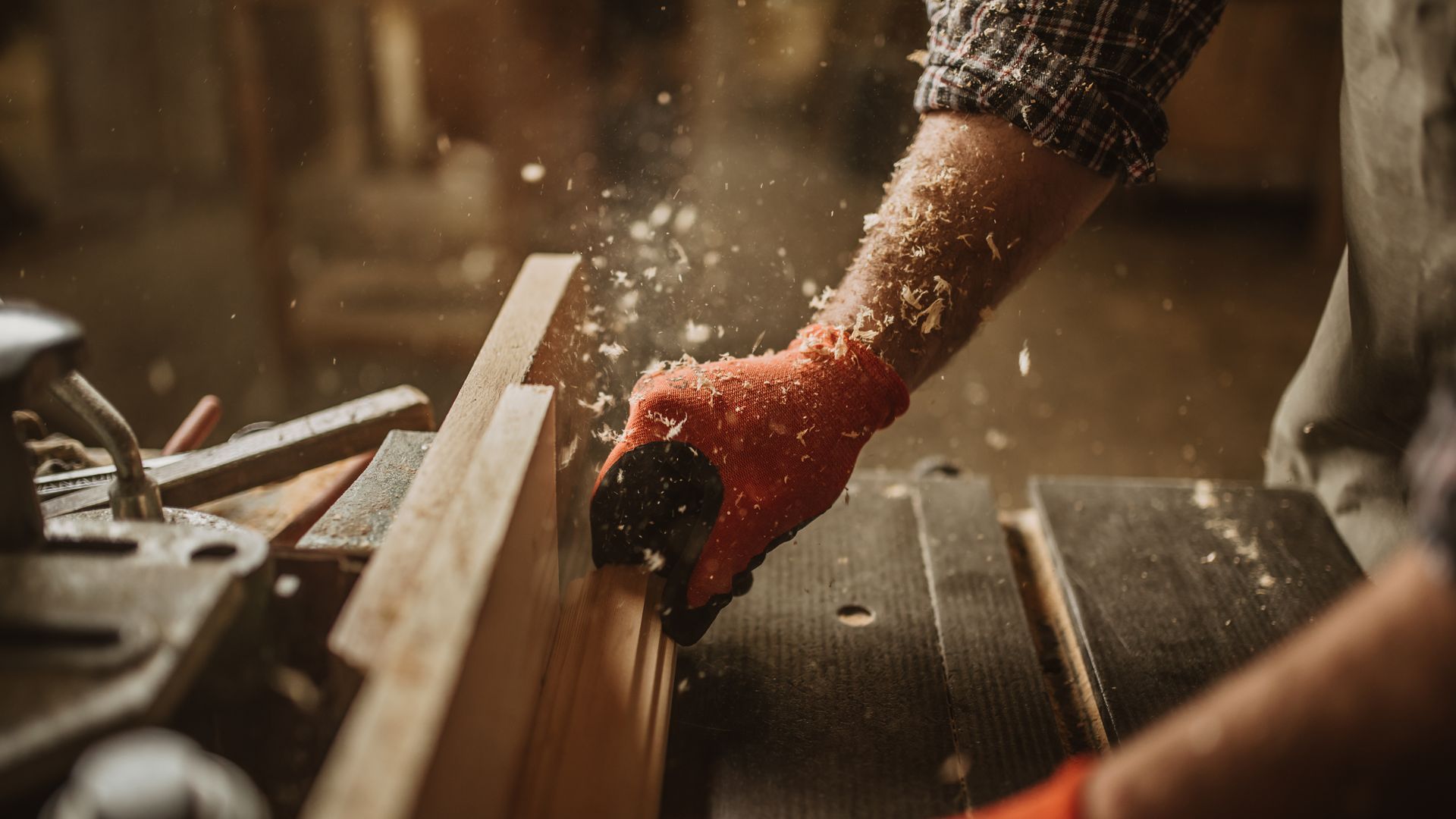 Des mains gantées manient une scie à main sur une planche de bois dans un atelier, de la sciure vole.