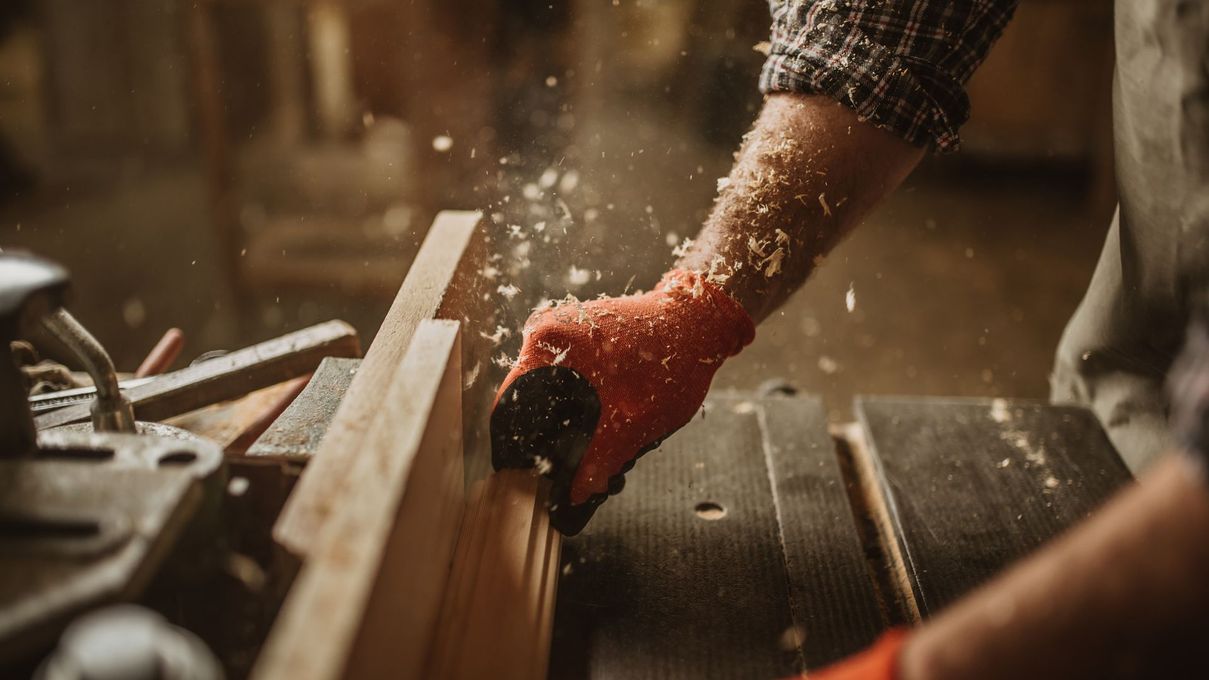 Des mains gantées poncent une planche de bois dans un atelier, de la sciure vole dans tous les sens.