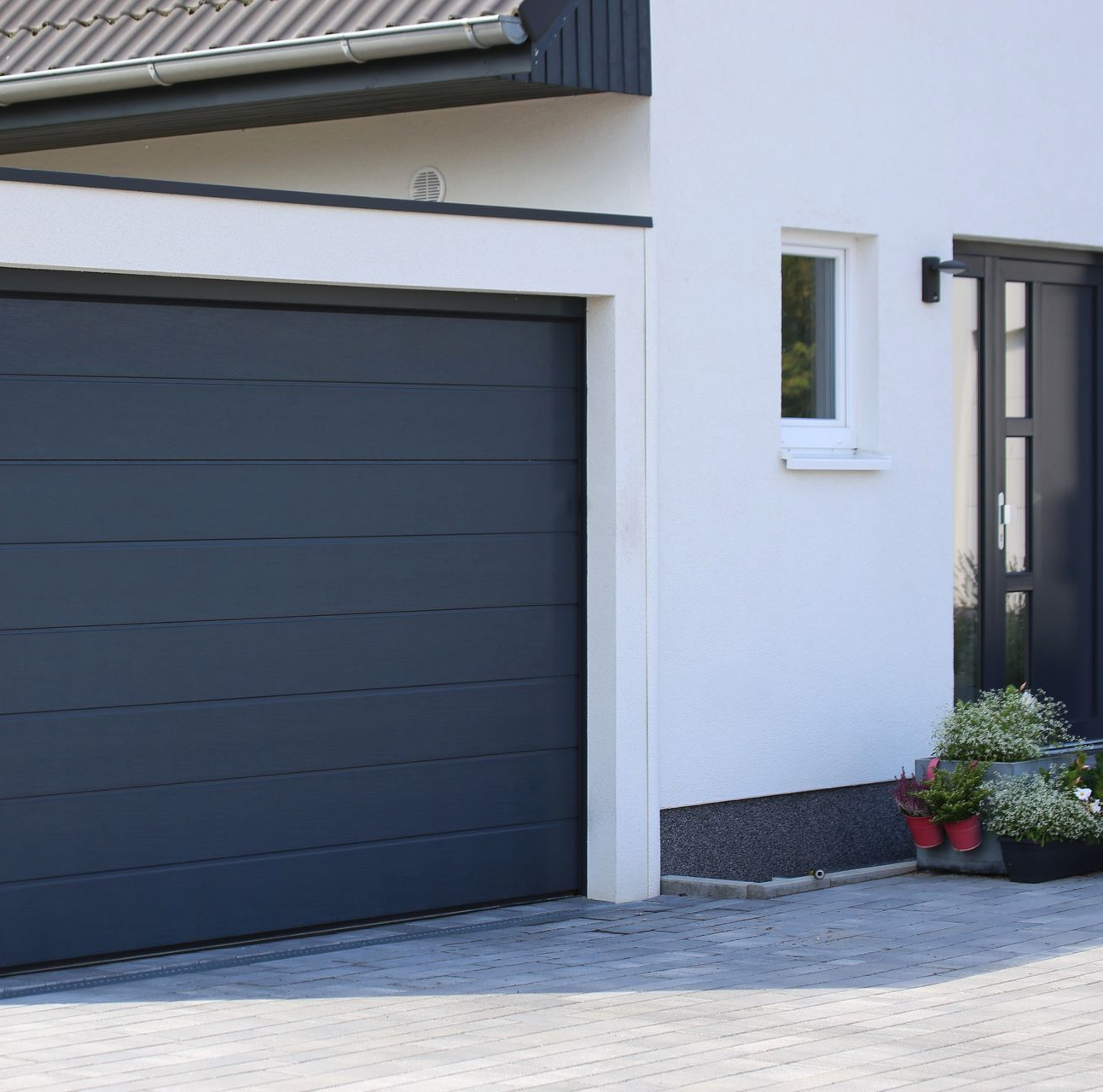 Extérieur d'une maison moderne avec une porte de garage foncée, des murs blancs et une petite plante en pot près de l'entrée.
