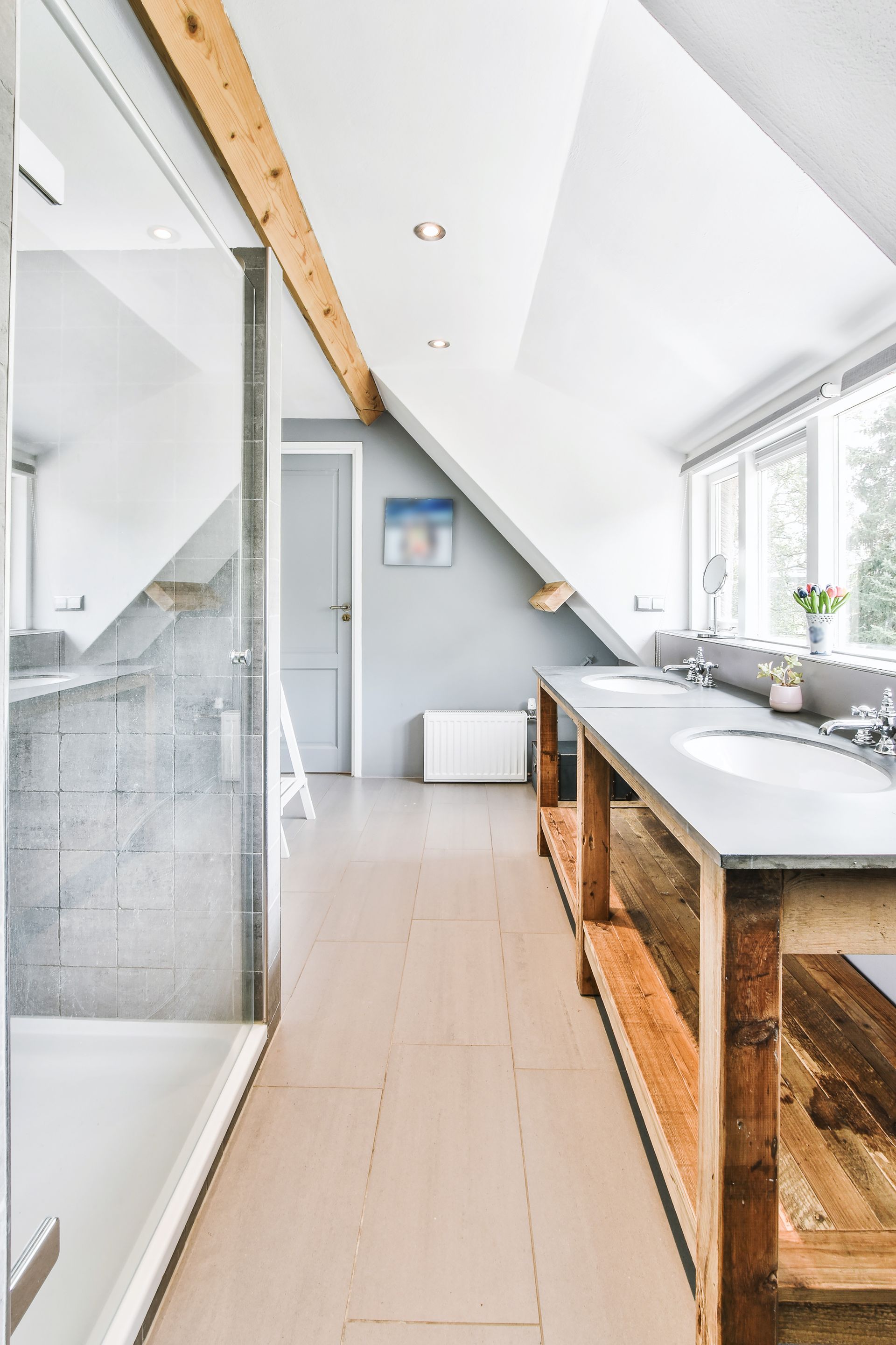 Salle de bains mansardée lumineuse avec plafond en pente, douche en verre et meuble-lavabo rustique près d'une fenêtre