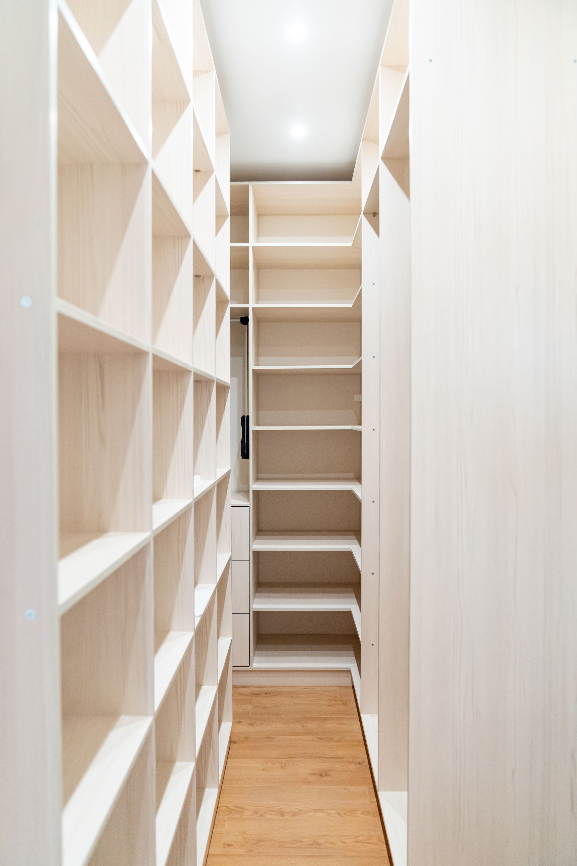 Couloir étroit et blanc avec étagères intégrées de chaque côté et parquet menant à un mur du fond lumineux.