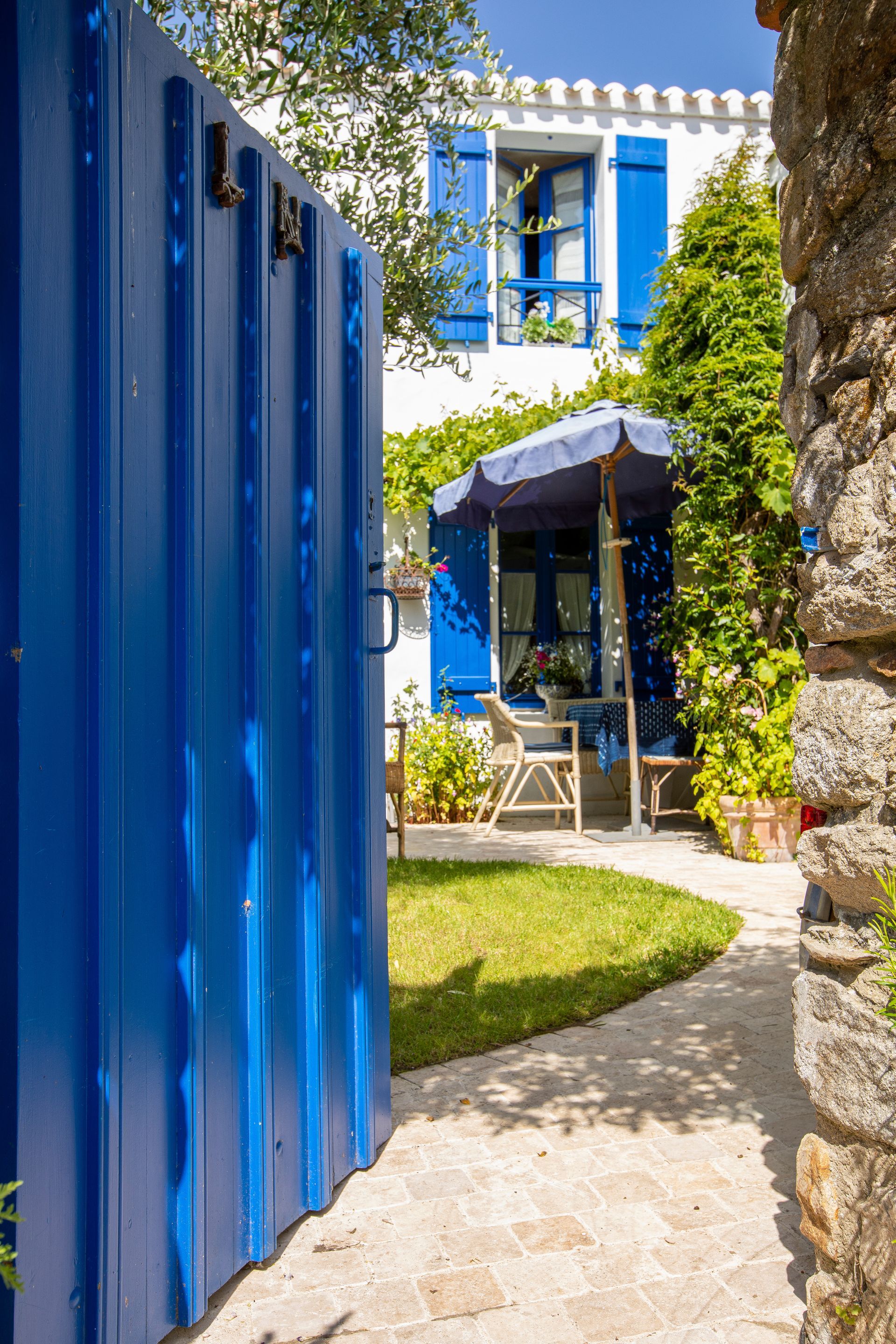 Un portail bleu s'ouvre sur une cour blanche et bleue avec un chemin de pierre, une pelouse verte et un parasol.