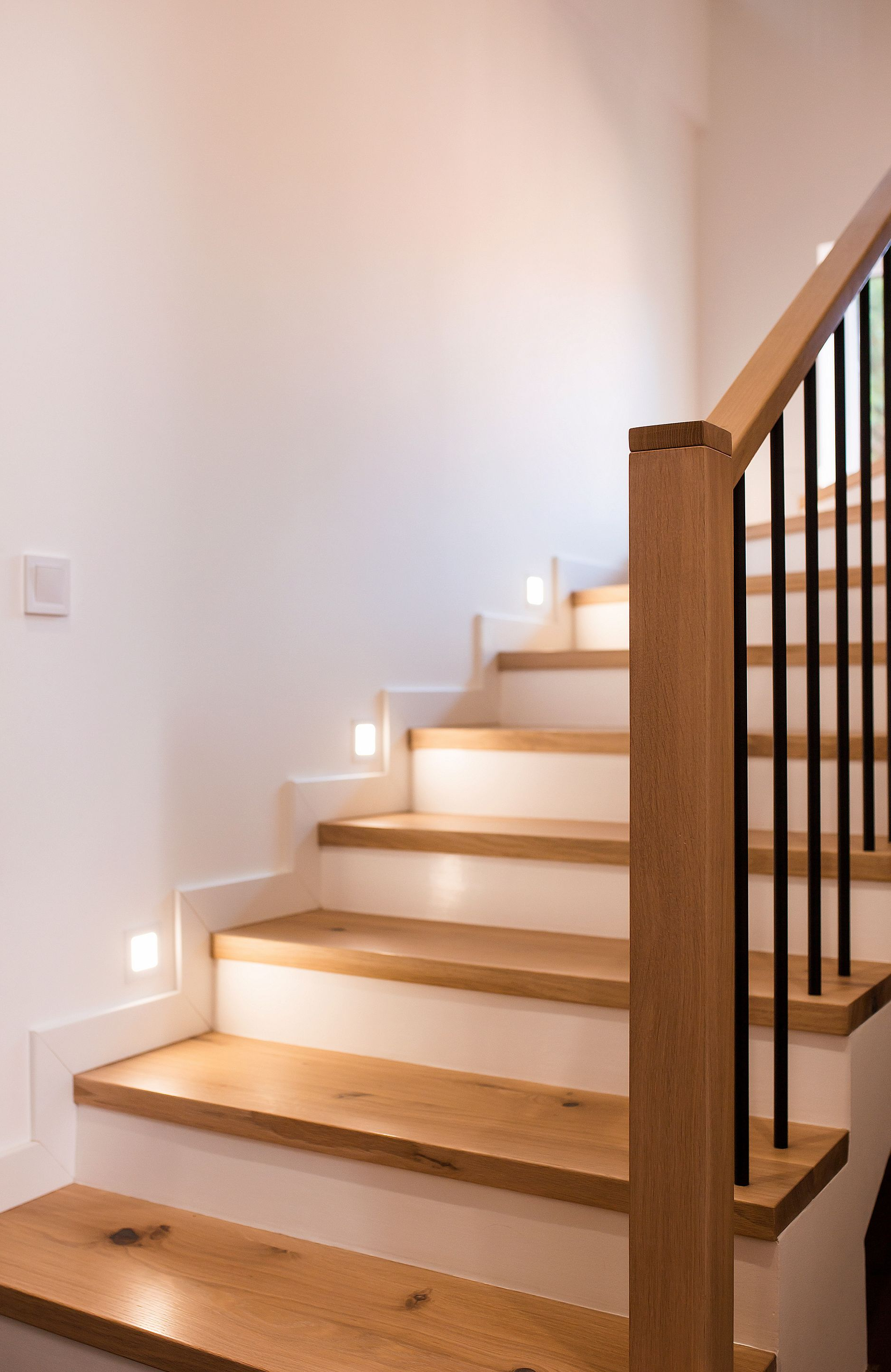 Un escalier en bois avec une rampe noire se trouve dans un couloir blanc lumineux, éclairé par de petites lampes de marche.