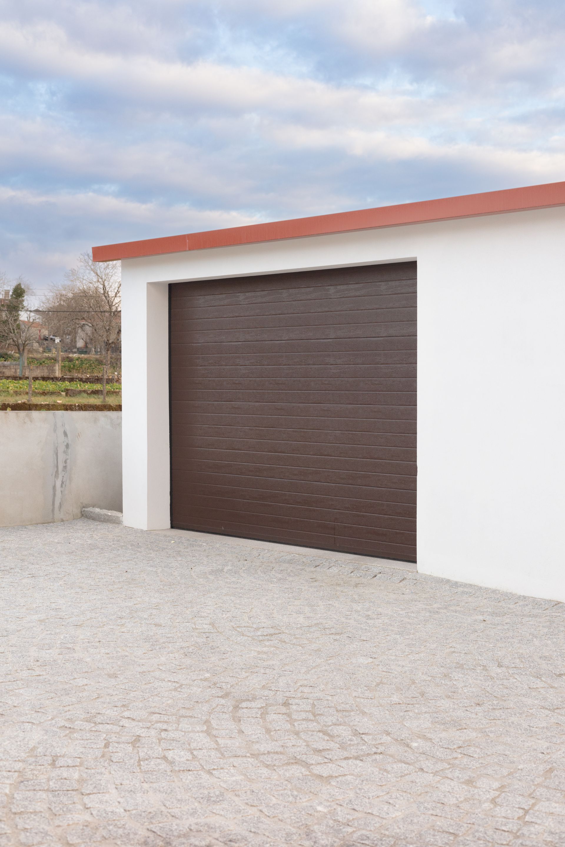 Garage blanc avec porte marron fermée et toit rouge sur une allée de gravier.