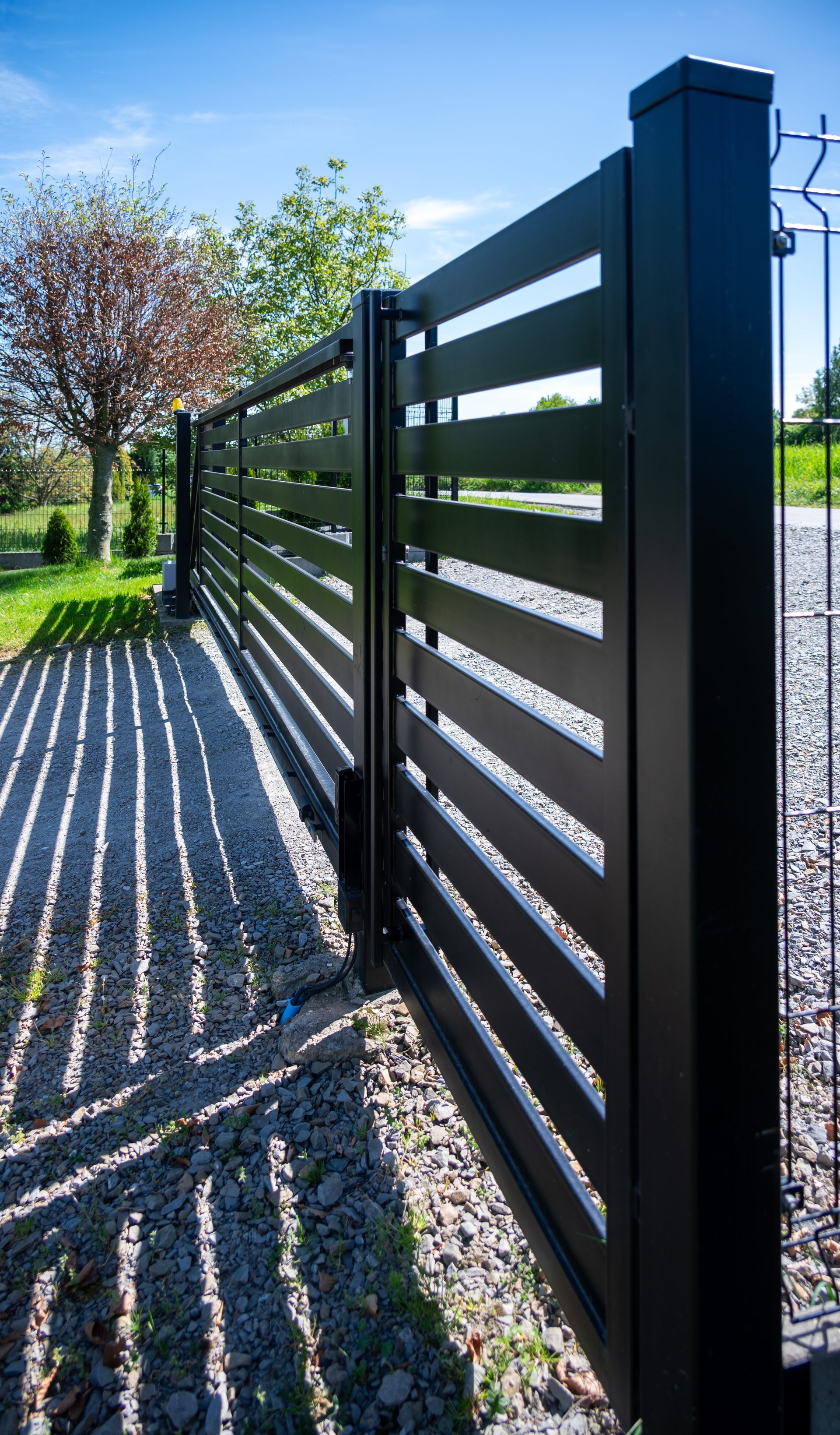 Une clôture en métal noir projette des ombres rayées le long d'un chemin de gravier ensoleillé dans un jardin verdoyant.