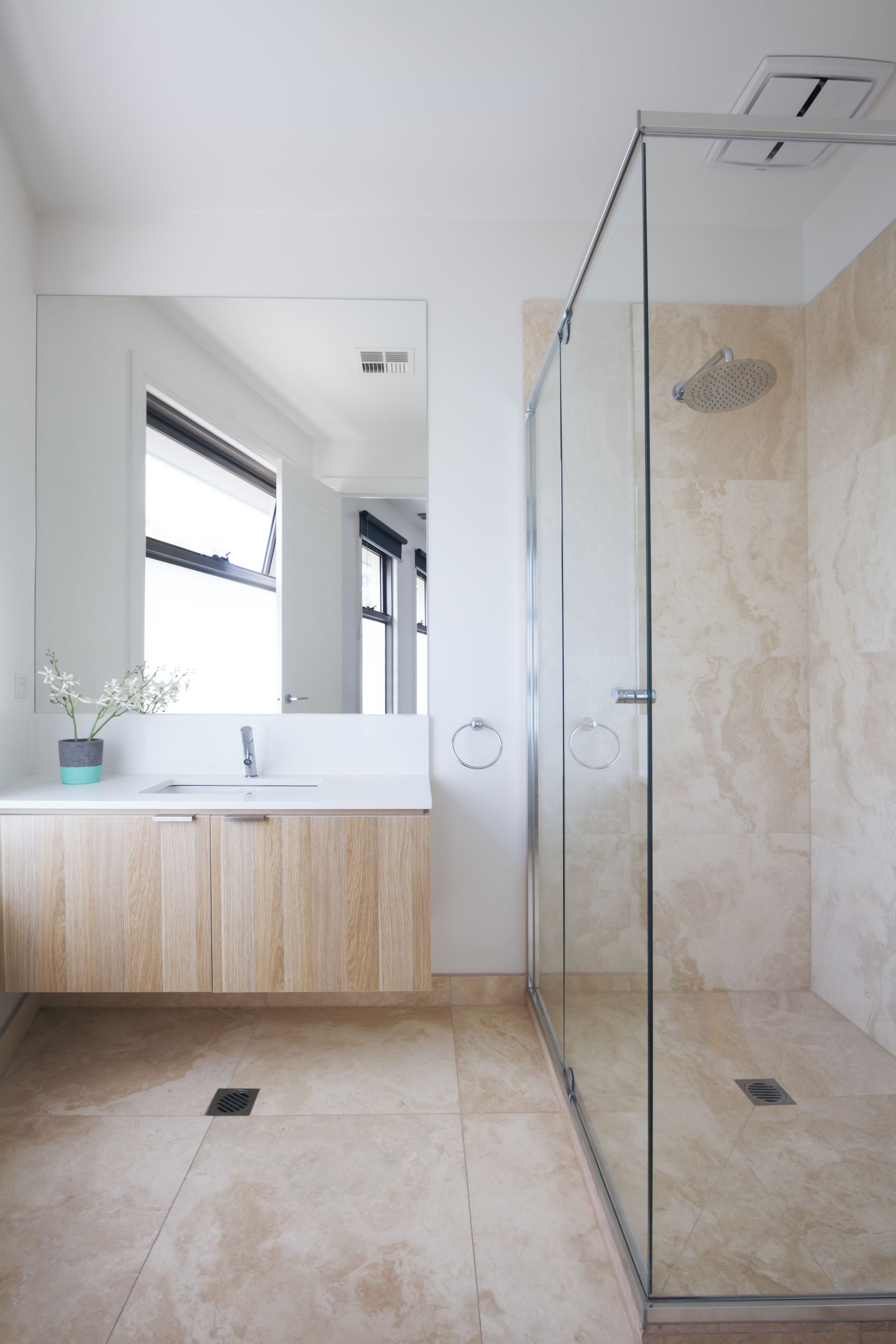 Salle de bains moderne avec douche en verre, miroir mural, meuble-lavabo suspendu en bois et carrelage beige au sol.