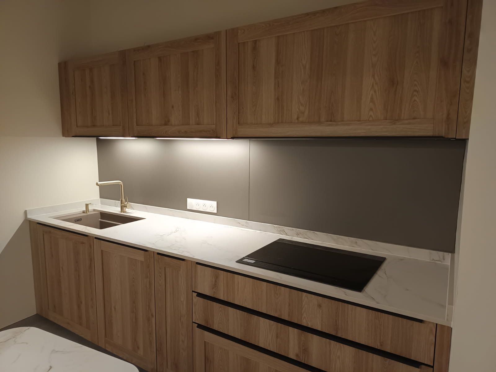 Petite cuisine avec placards en bois, plans de travail clairs et plaque de cuisson foncée.