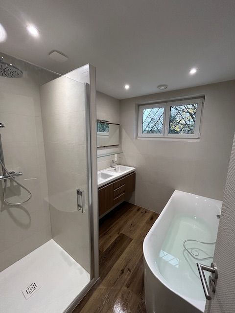 Salle de bains avec douche, meuble lavabo et baignoire. Parquet clair, murs aux tons neutres et fenêtre.