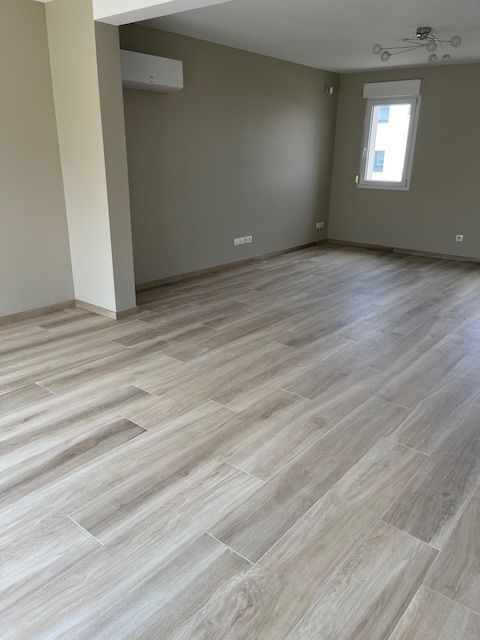 Pièce vide avec un sol imitation bois, des murs gris clair et une fenêtre.