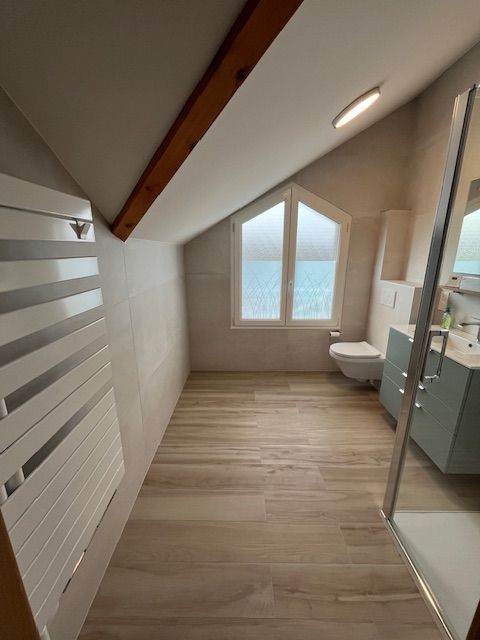 Salle de bains avec parquet clair, plafond mansardé, murs blancs et fenêtre. Comprend des toilettes et une douche.