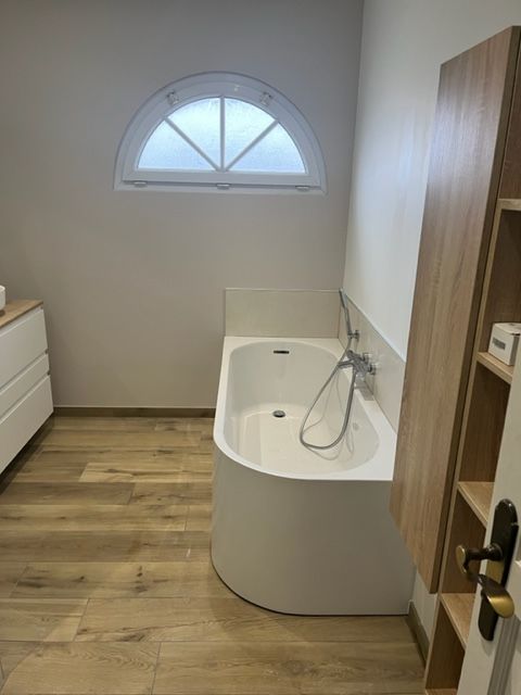 Petite salle de bains avec une baignoire incurvée sous une fenêtre cintrée et des étagères en bois.