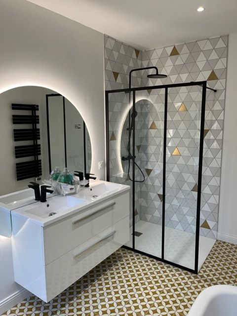 Salle de bains moderne avec douche à cadre noir, meuble-lavabo suspendu, miroir rond et carrelage géométrique au sol et aux murs.