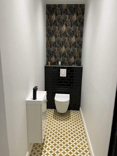 Petite salle de bains moderne avec sol carrelé à motifs dorés, toilettes noires, lavabo blanc et papier peint à motifs.