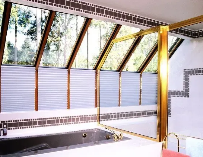 Banheiro com claraboia inclinada, banheira, acessórios dourados e persianas parcialmente fechadas.