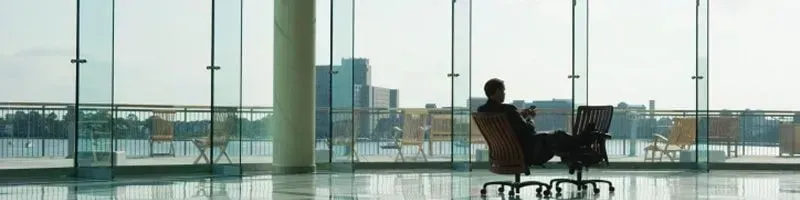 Uma pessoa em silhueta está sentada em uma cadeira perto de uma janela, olhando para um laptop, com vista para a cidade.