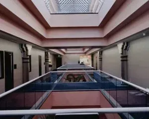 Corredor com seções de piso de vidro e teto com claraboia. Paredes e pilares bege, com detalhes em rosa.