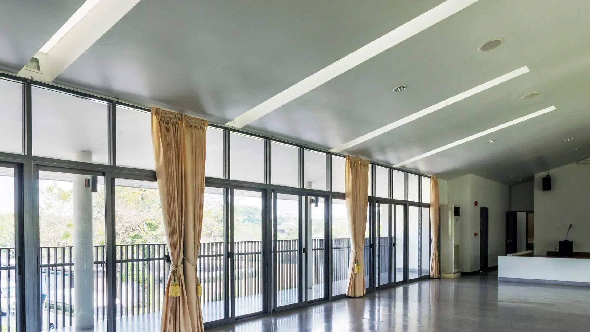 Espaço interior luminoso com grandes janelas de vidro e cortinas bege com vista para uma varanda.