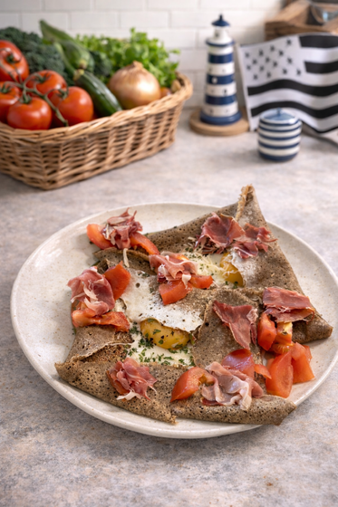 Une crêpe de sarrasin garnie de prosciutto, de tomates et d'un œuf au plat, servie sur une assiette blanche, avec un panier de légumes derrière.