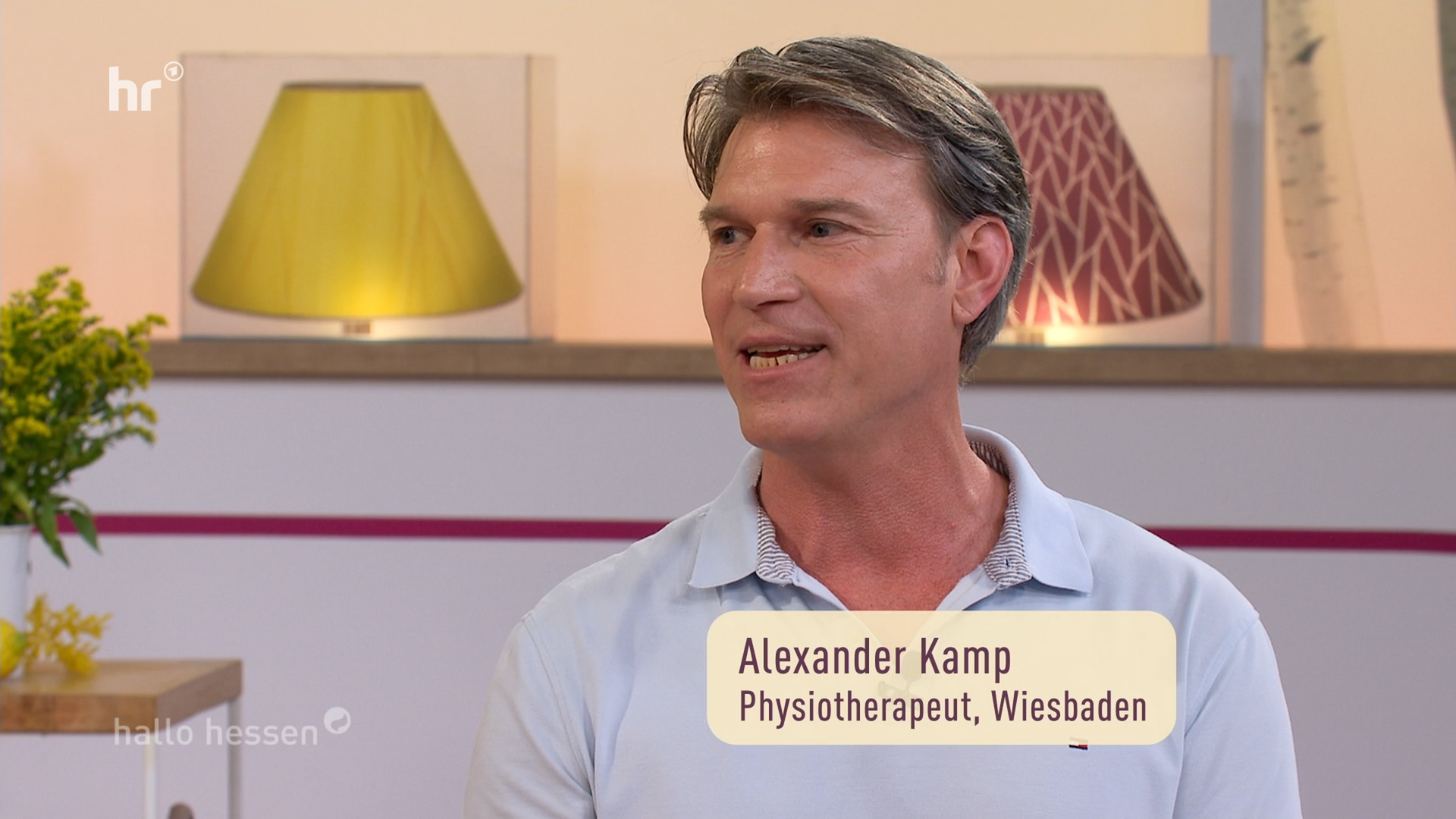 Ein Mann in einem grauen Polohemd spricht in einem hellen Studio mit Lampen und einer Topfpflanze; im unteren Drittel steht „Alexander Kamp“.