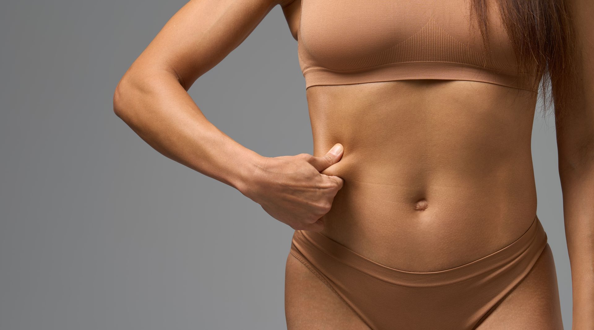 Une femme se pince la peau du ventre, vêtue d'un crop top beige et d'un sous-vêtement, sur fond gris.