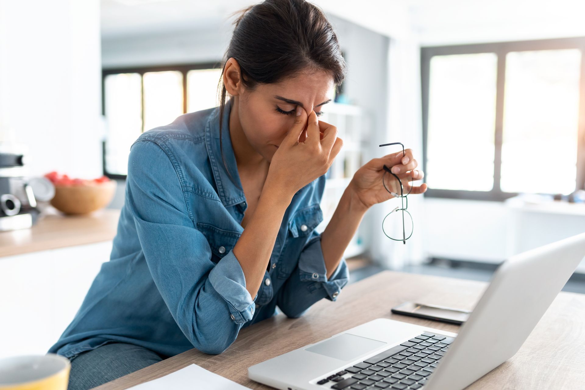 Femme à l'air fatigué, tenant des lunettes, assise devant son ordinateur portable dans la cuisine.