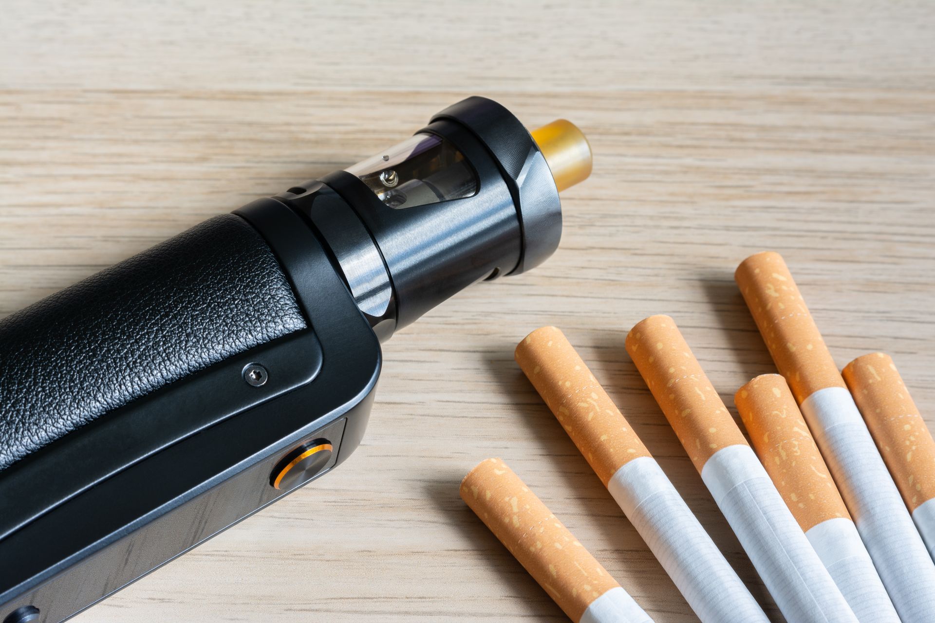 Une cigarette électronique noire à côté de plusieurs cigarettes sur une surface en bois clair.