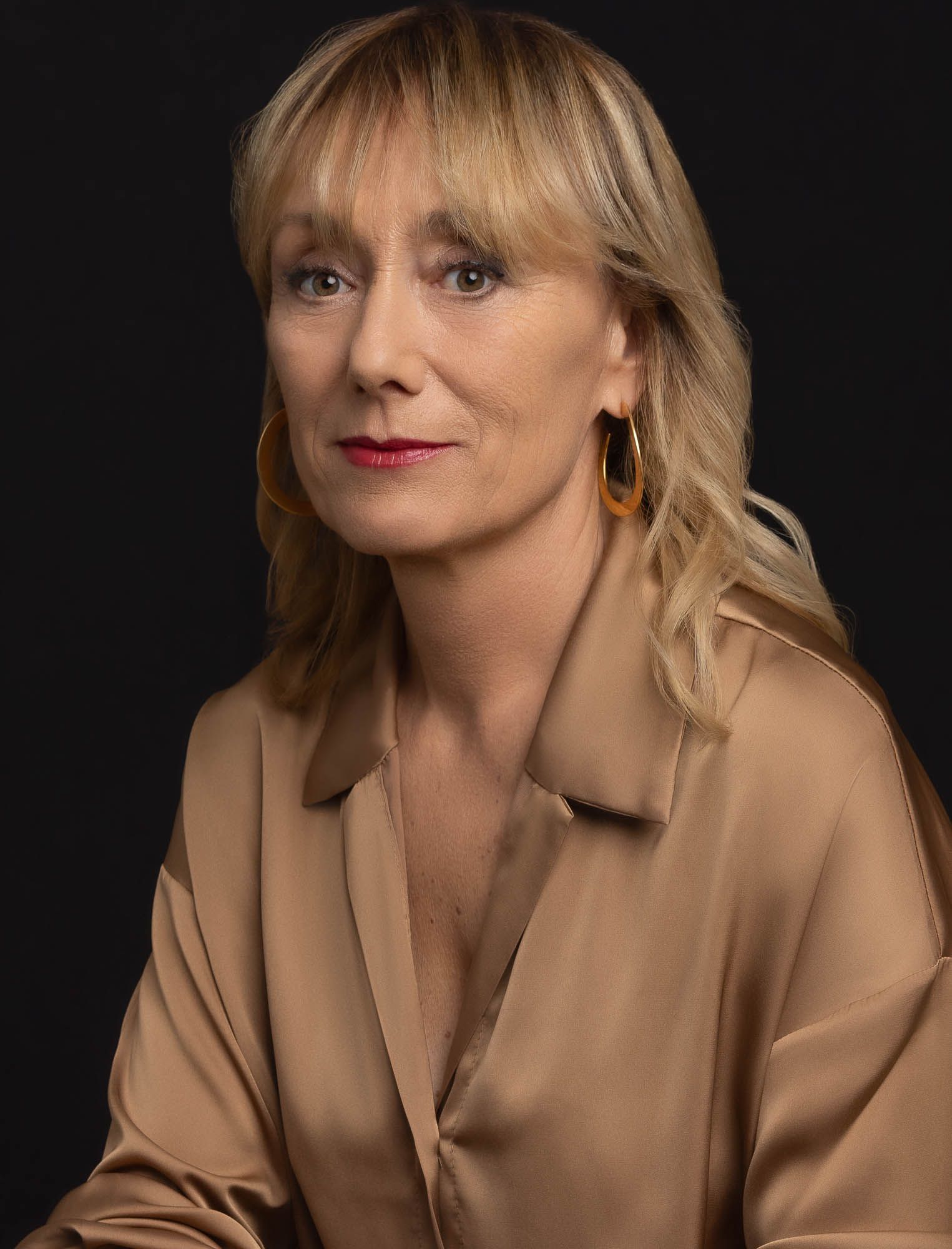 Femme aux cheveux blonds, vêtue d'une chemise en satin beige et de boucles d'oreilles créoles dorées, regardant l'objectif.