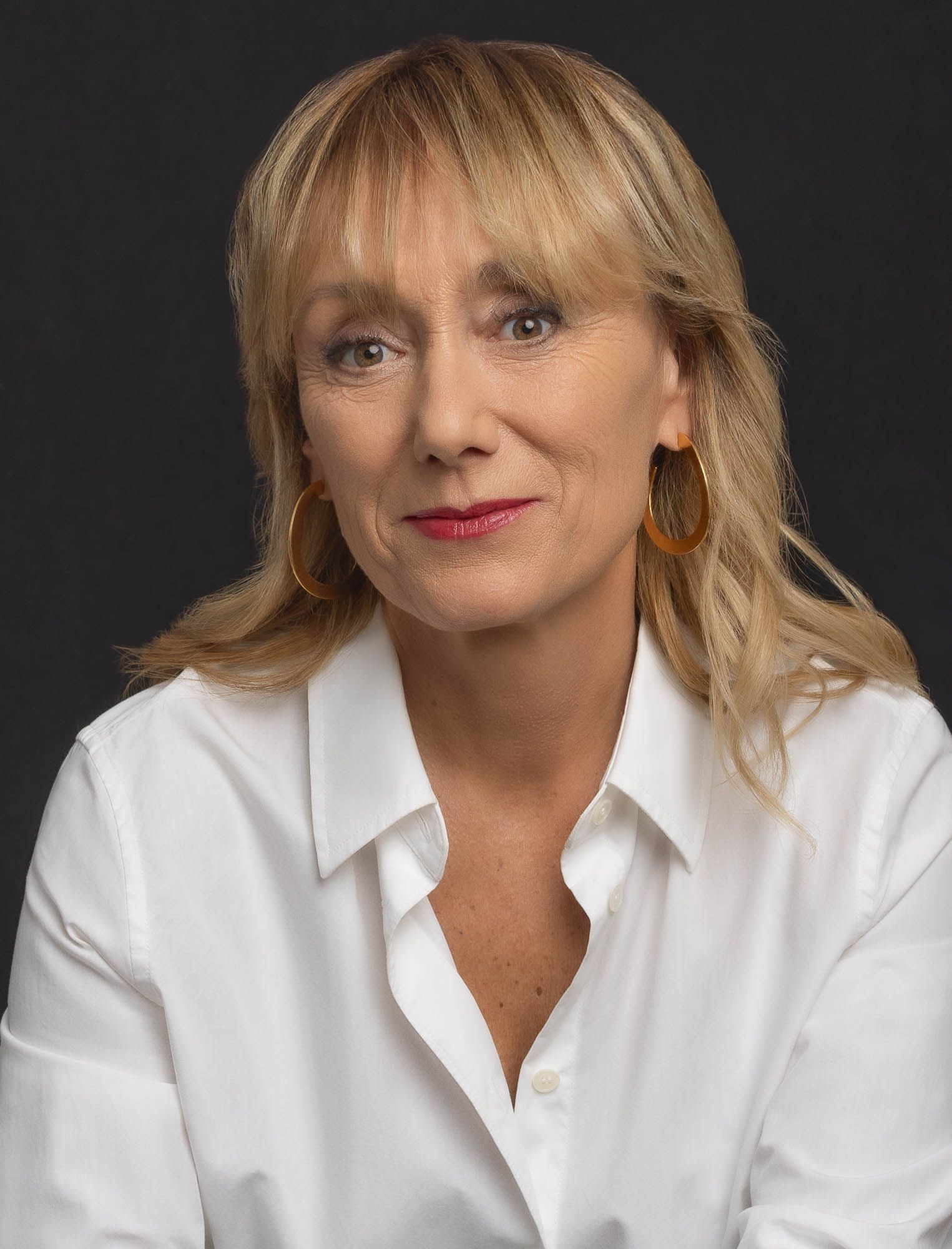 Femme aux cheveux blonds, portant une chemise blanche et des boucles d'oreilles créoles dorées, sur fond noir.