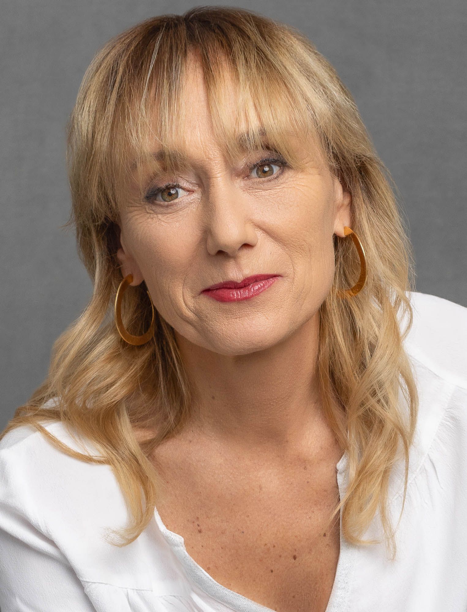 Femme aux cheveux blonds ondulés, portant des créoles et une chemise blanche.